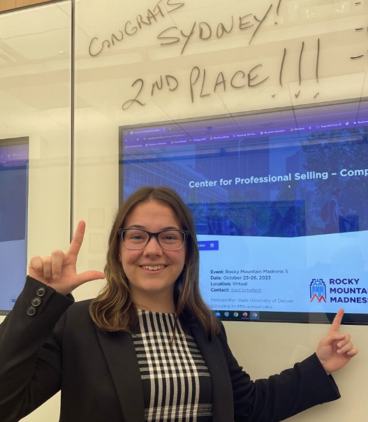 Woman with glasses and black jacket holds up an "L" with fingers and points to a computer screen. 