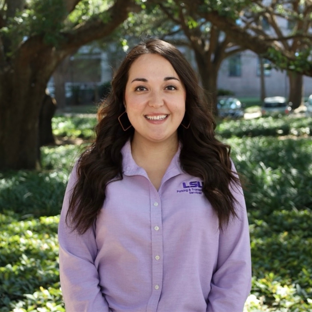 Woman in purple shirt smiling and standing in front of a tree and bushes